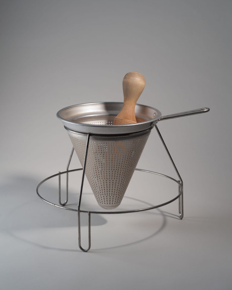 Conical sieve set into a circular wire frame with three legs and a handle. A wooden pestle sits inside the sieve.