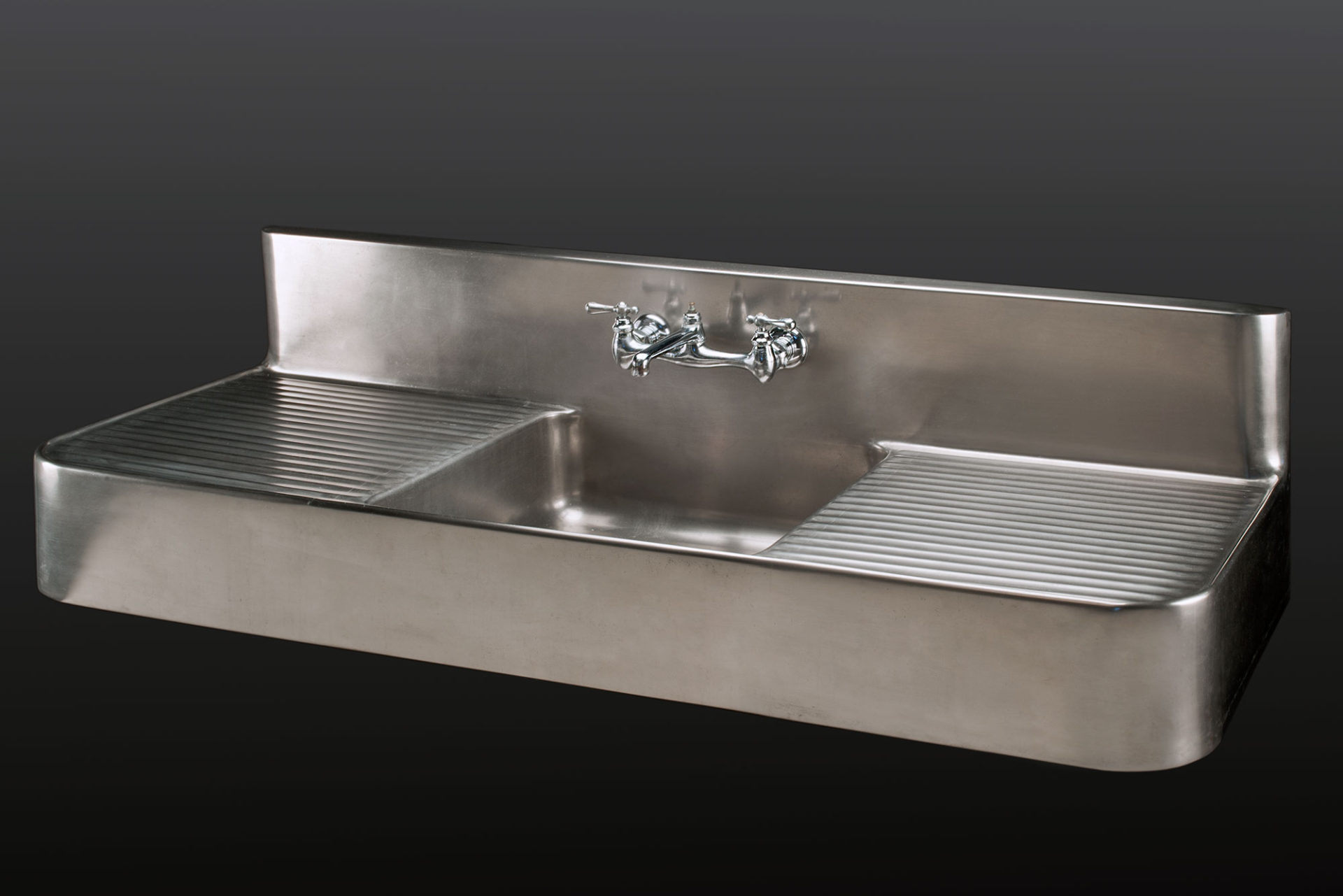 Large évier en métal avec plateaux de drainage de chaque côté de la cuve rectangulaire; poignées et robinets au centre du dosseret, lui aussi rectangulaire.