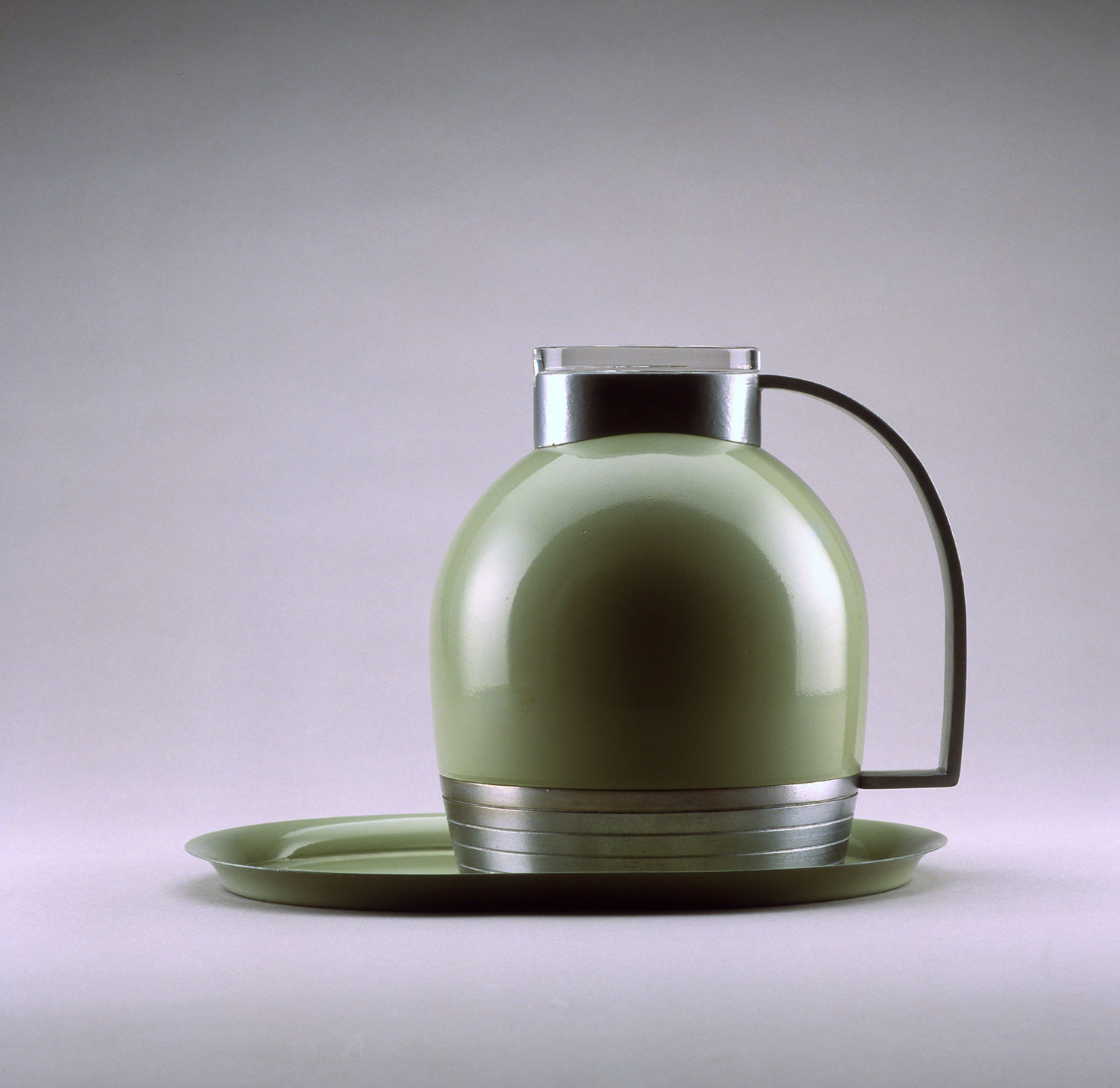 Dome-shaped thermal pitcher in green enameled metal with an aluminum base, top, and handle, and a lid of clear glass. It sits on an oval tray in matching green.
