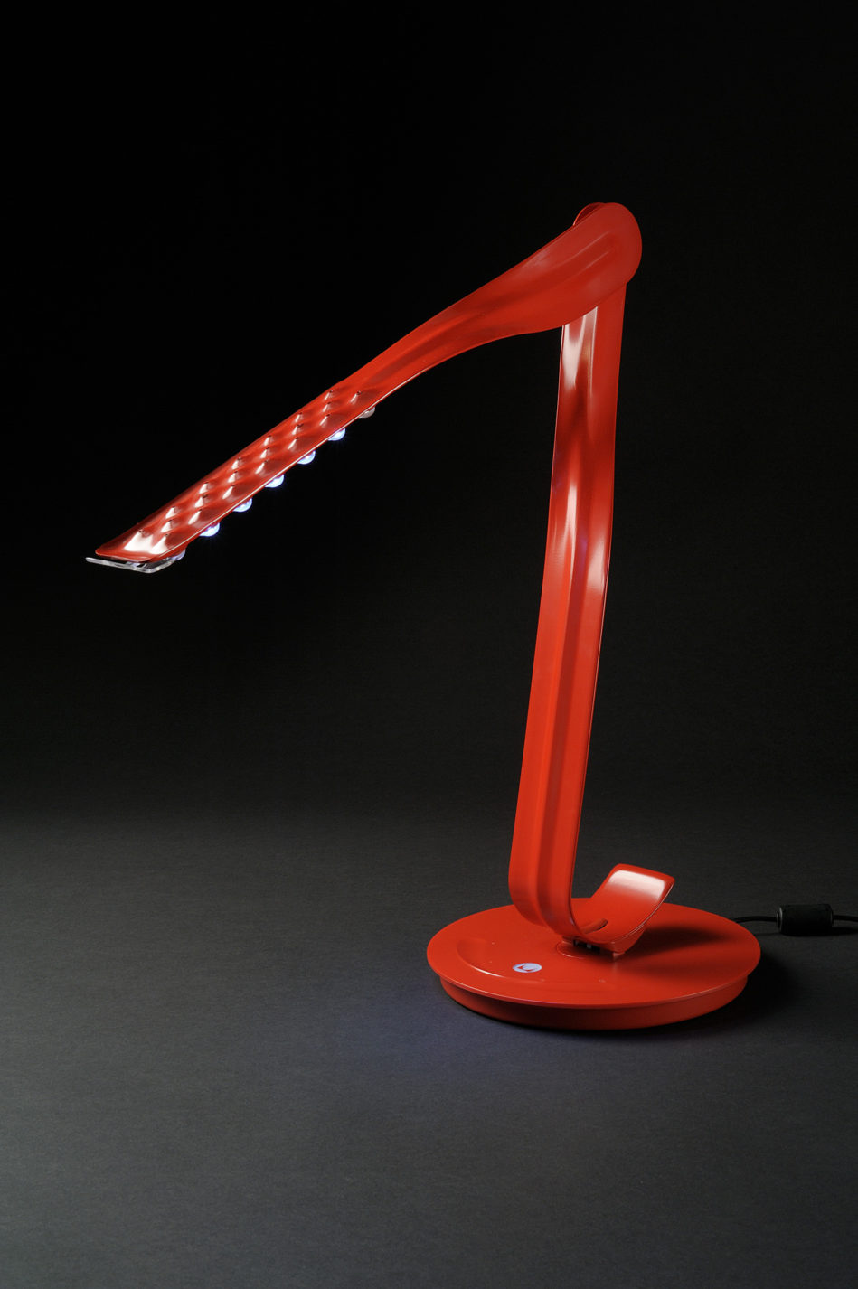 Bright red LED lamp. Two bent strips of metal connect at a movable joint to form the body and arm above a circular base.