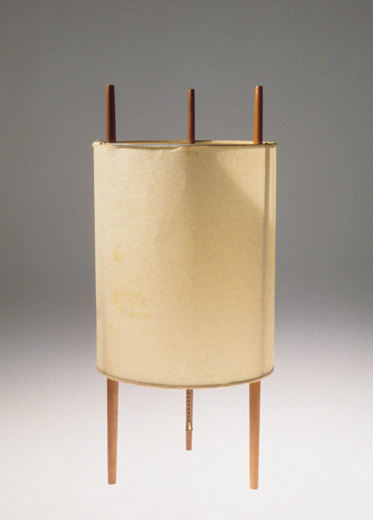 Lampe composée de trois bâtons de bois verticaux entourés d’un abat-jour cylindrique.