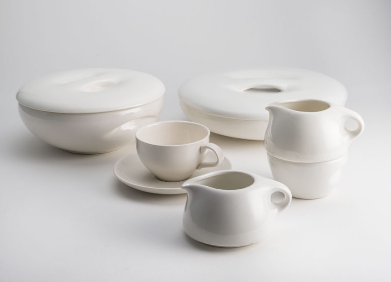 Set of white ceramic dishes. Two bowls, one shallower than the other, each have lids with indentations for handles. One stacking sugar bowl and creamer; one creamer, and one teacup and saucer.