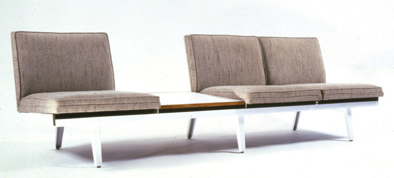 Armless sofa with three seats with tan seat and back cushions on a low steel bench. Between two of the seats is a white Formica table surface.
