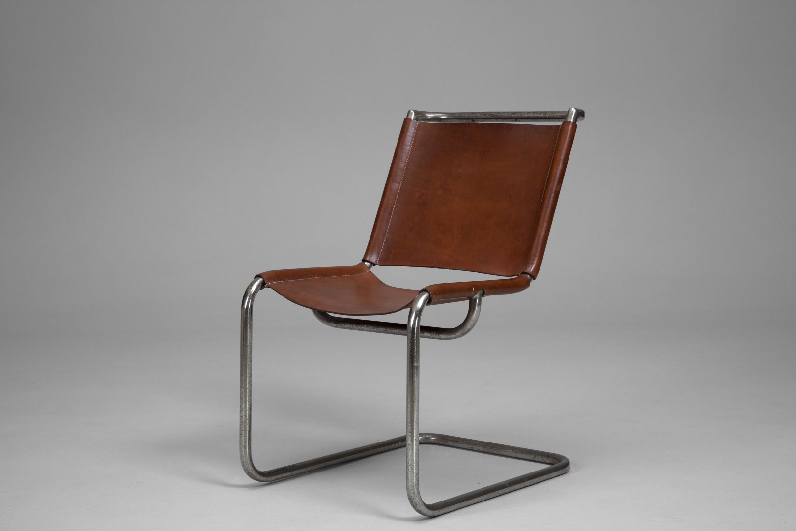 Cantilevered chair with tubular metal frame and a sheet of brown leather suspended between the bars for the seat and another for the back.