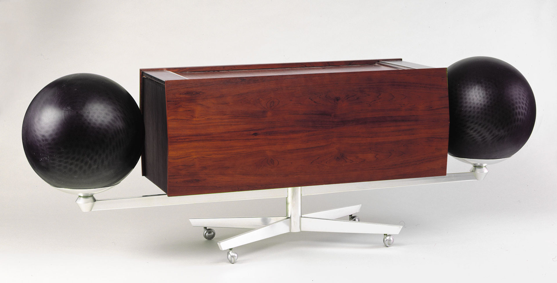 Rectangular wooden cabinet with a large black spherical speaker at each end, mounted on an angular aluminum base.