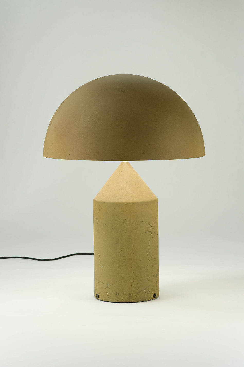 Lampe de table beige à base cylindrique dont l’extrémité forme un cône, coiffée d’un abat-jour demi-sphérique opaque.