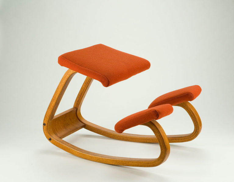 Chaise à appuie-genoux composée d’une longue pièce de bois courbé; siège et appuie-genoux coussinés et recouverts de tissu orange.