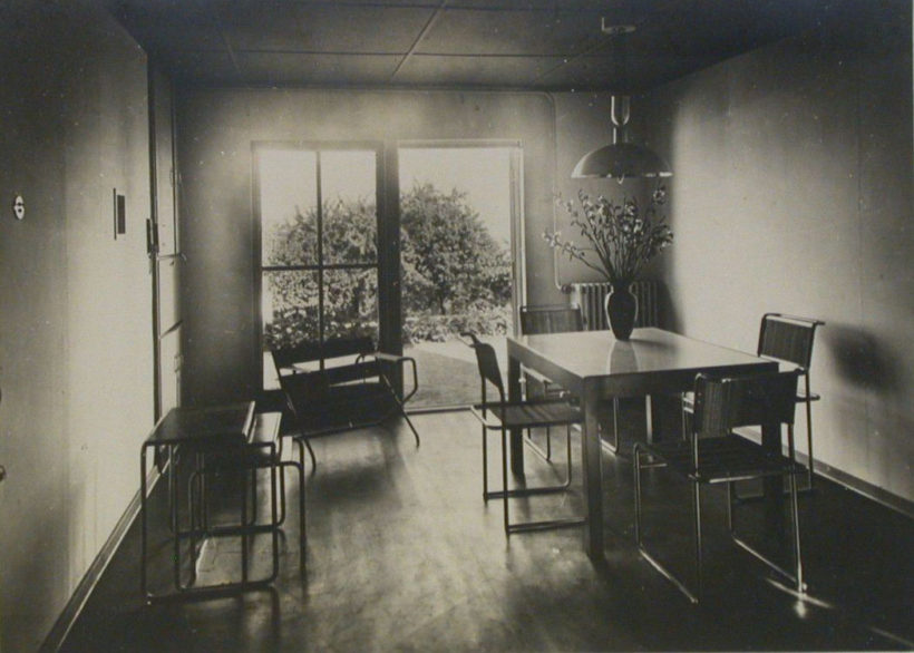 Black-and-white interior view showing a minimalistic arrangement of modern furniture, including dining table with B5 chairs and a set of nesting tables.