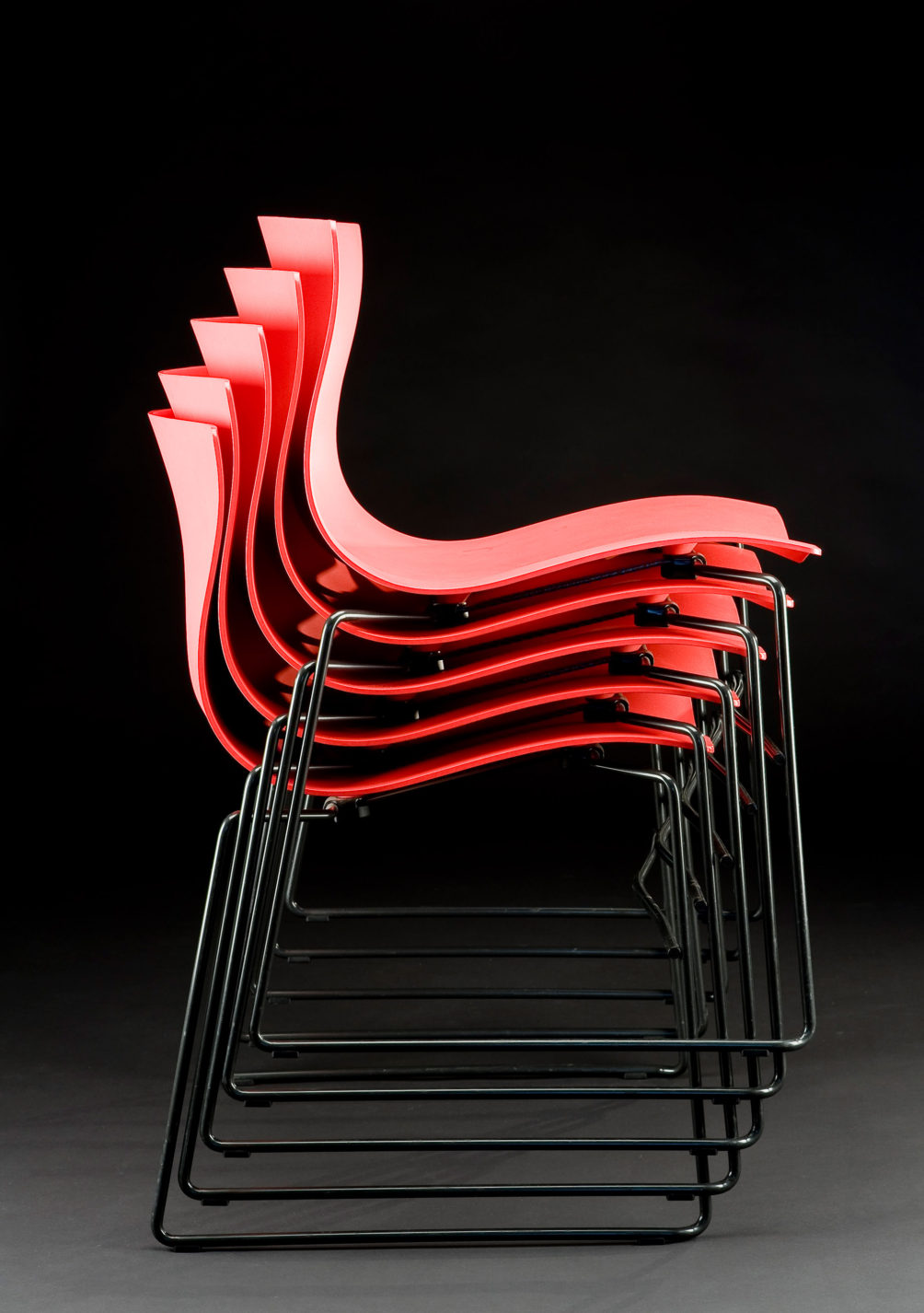 Stack of five chairs, each with undulating shell seat and back in red plastic supported black tubular-steel legs.