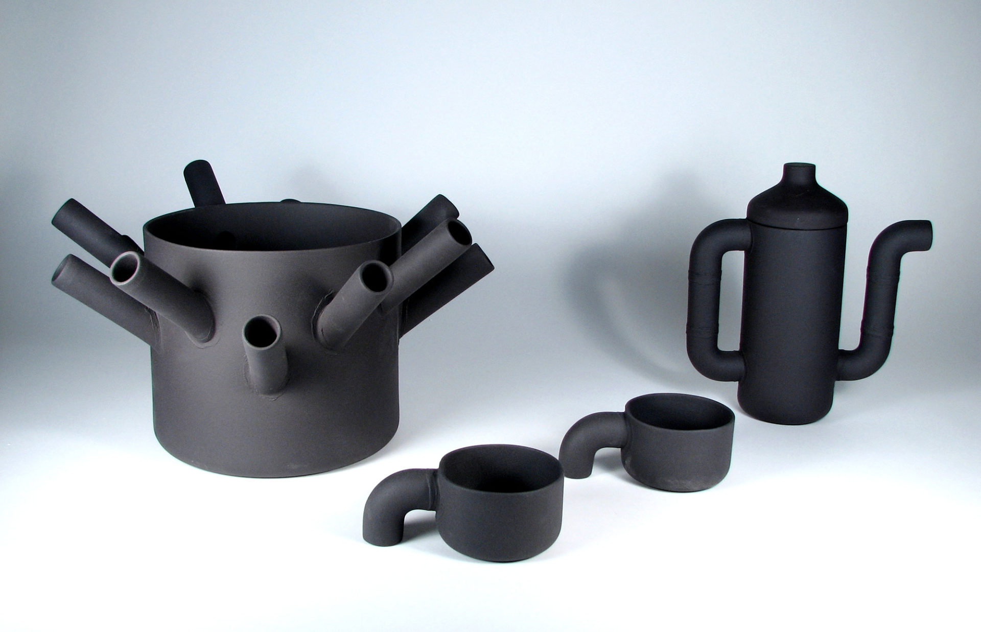Vase, cafetière et tasses gris foncé avec éléments rappelant des pièces de plomberie.